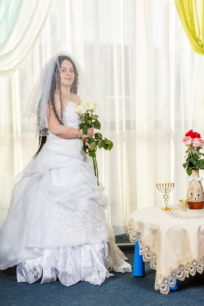 Een vrolijke Joodse bruid met haar gezicht bedekt met een sluier en een boeket witte rozen staat in de synagoge voordat ze het Huppa-ritueel uitvoert aan een tafel met bloemen. Verticale foto