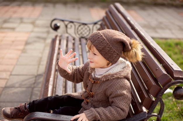 een vrolijke kleine blonde jongen in gebreide bruine kleren zit op een bankje in het park