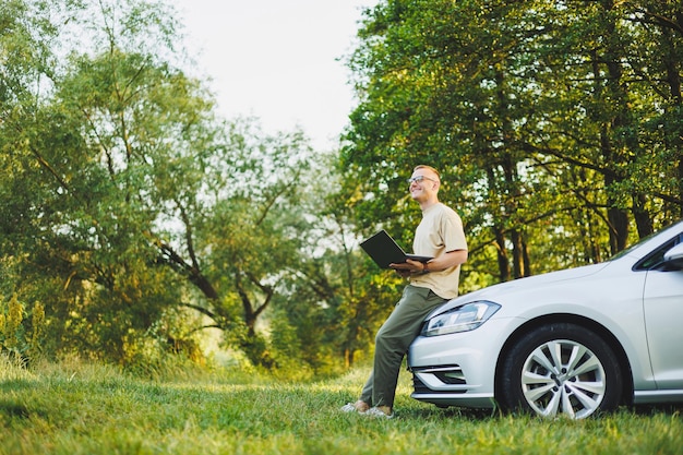 Een vrolijke man met een bril zit op de motorkap van zijn auto in de natuur en werkt op een laptop Freelance werk op afstand tijdens vakantie