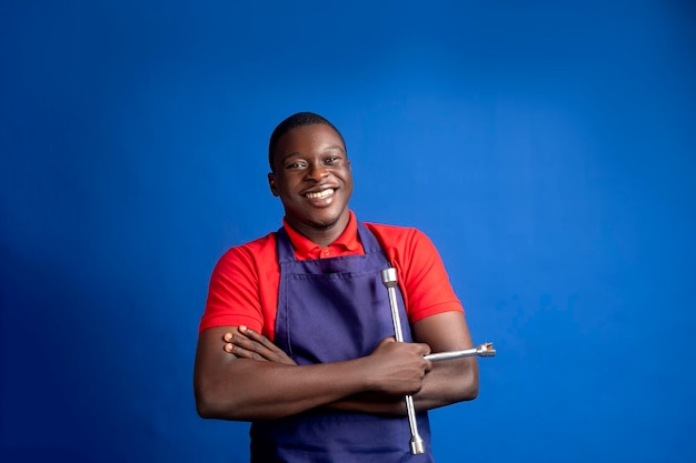 Een vrolijke man met een rood shirt en een blauwe schort houdt met vertrouwen gereedschappen vast tegen een blauwe achtergrond