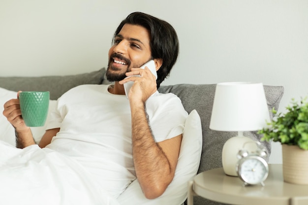 Een vrolijke man uit het Midden-Oosten zit op witte bedoproepen via de telefoon en communiceert genieten van een kopje koffie