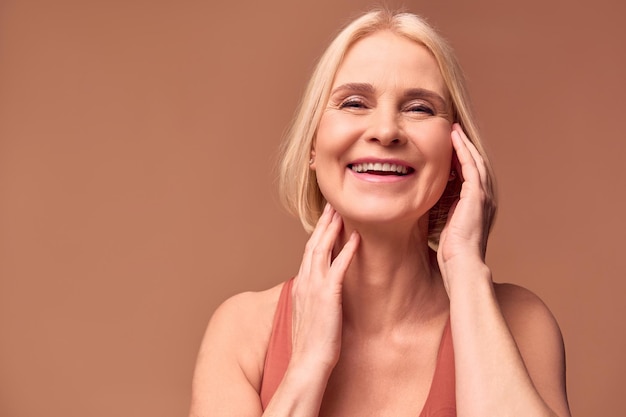 Foto een vrolijke oudere vrouw raakt de huid van haar gezicht aan met haar handen en lacht het concept huidverzorging vrouwelijke schoonheidbeige achtergrond