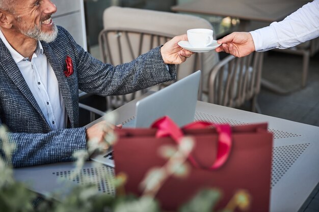 een vrolijke senior man die een kopje met een warme drank neemt van een ober in het café