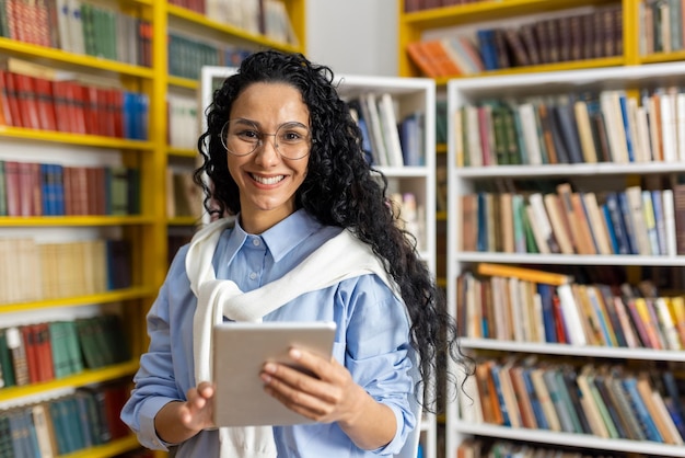 Foto een vrolijke student met een digitale tablet staat in een bibliotheek met een brede glimlach omringd door