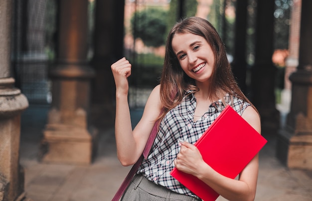 Een vrolijke student met een rood notitieboekje in de buurt van de campus slaagde voor het examen en zei ja