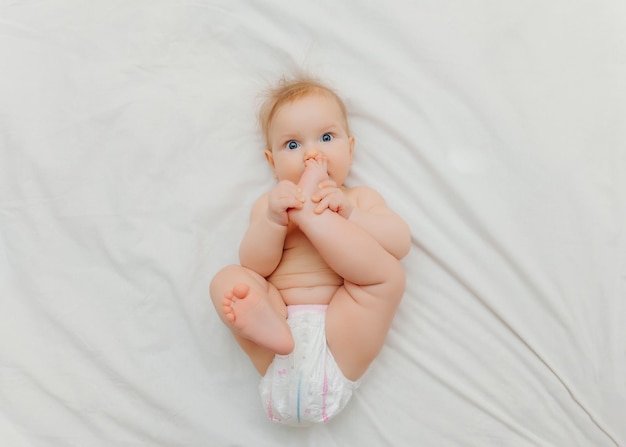 Een vrolijke, vrolijke baby in luiers ligt op een wit bed en houdt zijn been vast Hoogwaardige fotoplaats voor tekst