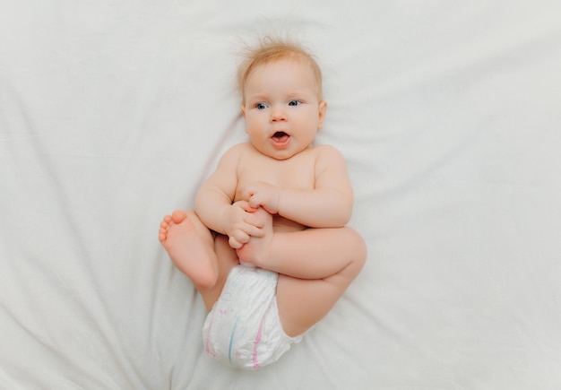 Een vrolijke, vrolijke baby in luiers ligt op een wit bed en houdt zijn been vast Hoogwaardige fotoplaats voor tekst