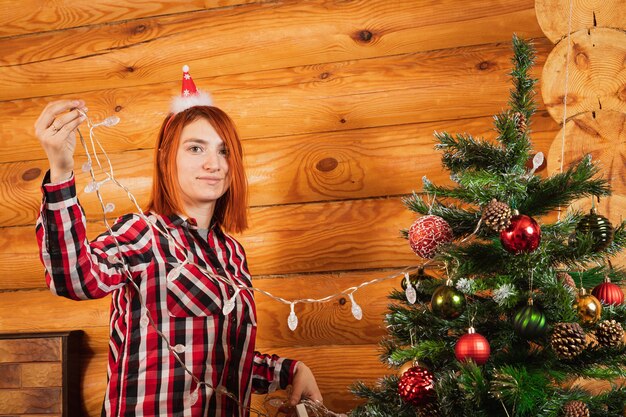 Een vrolijke vrouw in een geruit overhemd hangt een krans aan een kerstboom op de achtergrond van een brandende open haard