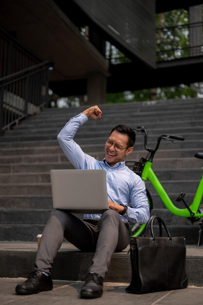 Een vrolijke zakenman zit op de trappen en werkt aan een laptop met een glimlach en juicht met een opgeheven vuist