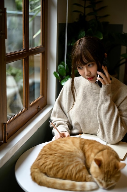 Een vrouw aan de telefoon terwijl haar kat op een tafel ligt te slapen