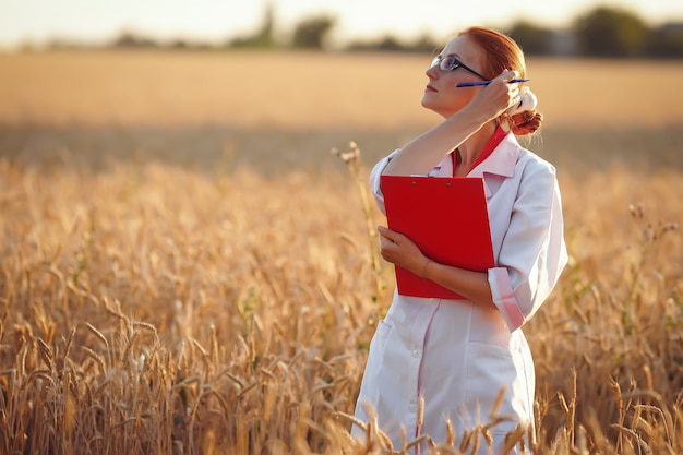 een vrouw agronoom in een veld van tarwe met een rode map en een pen in witte jassen en glazen bij de oogst bij zonsondergang