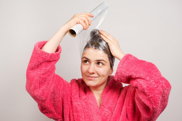 Een vrouw brengt een masker aan op haar haar en wikkelt een film om haar hoofd glimlachend in een roze badjas Het concept van haarverzorging