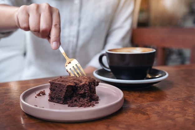 Een vrouw brownie cake met vork met koffiekopje snijden op houten tafel in café