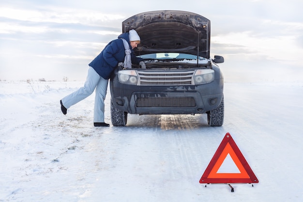 Een vrouw die dichtbij de kapotte auto naar de motor kijkt. rond de winter en sneeuwveld. er is een noodsituatiebord.