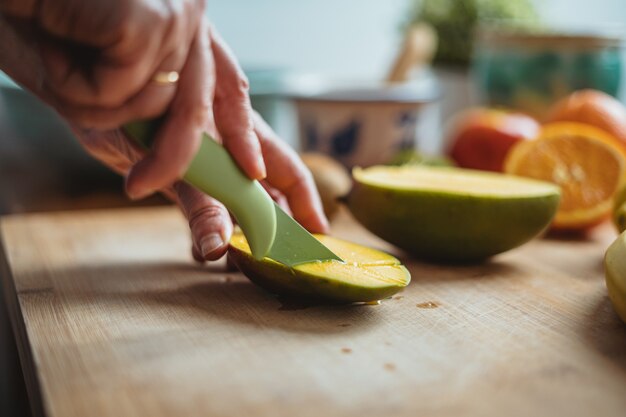 Een vrouw die een mango snijdt met een groen mes op een houten tafel omringd door ander fruit.