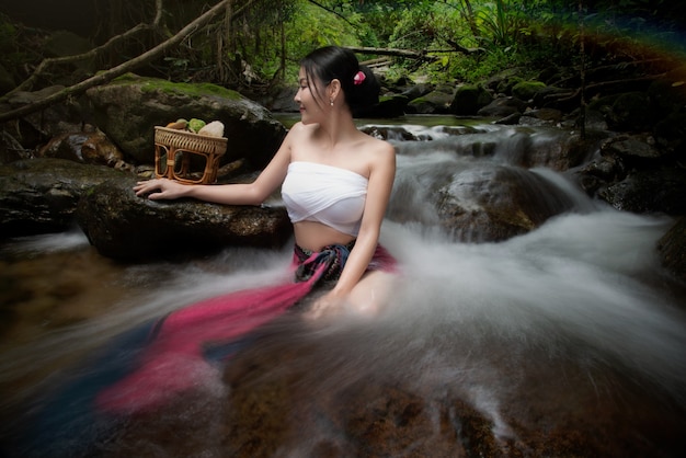 Foto een vrouw die een traditionele lanna thai-jurk en traditionele spa-apparatuur draagt, zit bij een waterval.