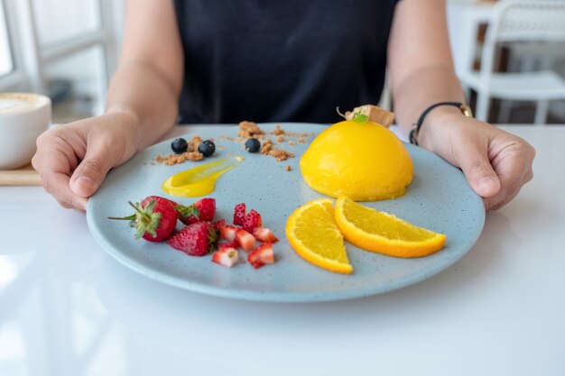 Een vrouw die en een plaat van oranje cake met gemengd fruit in koffie houdt toont