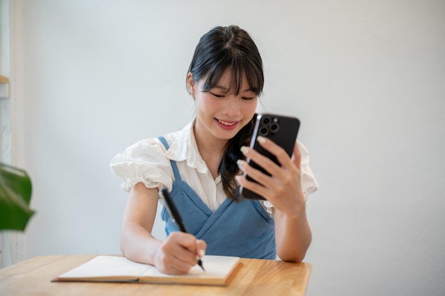 Een vrouw die lijstjes maakt in haar notitieboekje terwijl ze haar smartphone gebruikt terwijl ze binnenshuis aan een tafel zit