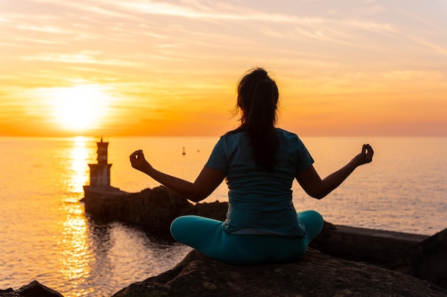 Een vrouw die meditatie doet op een rots bij zonsondergang naast een vuurtoren in de zee, gezond en naturistisch leven
