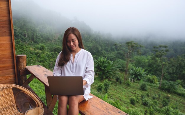 Een vrouw die op een mistige dag een laptop gebruikt en eraan werkt terwijl ze op het balkon zit met een prachtig uitzicht op de natuur