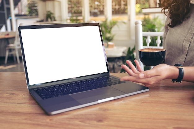 Een vrouw die werk met laptop presenteert met een leeg wit scherm op houten tafel terwijl ze koffie drinkt