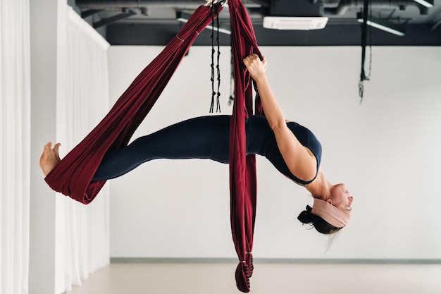 Een vrouw doet yoga op een hangende bordeauxrode hangmat in een lichte sportschool.