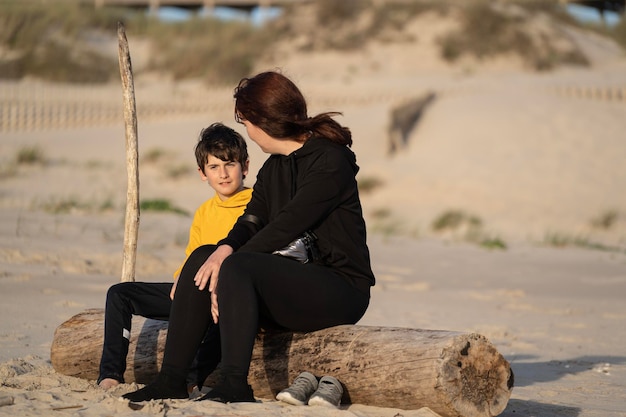 Een vrouw en een jongen zitten ontspannen op een boomstam op het strand en brengen samen tijd door