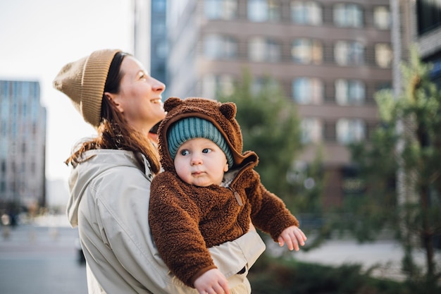 Een vrouw houdt een baby vast met een berenmuts en een bruin jasje.