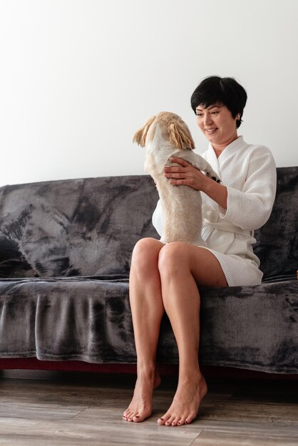 Een vrouw in een badjas die op de bank zit en speelt en haar hond knuffelt Brunette vrouw die thuis ontspant met haar kleine hond Liefde voor dieren Goedemorgenconcept Morgenroutine en huishoudelijke levensstijl