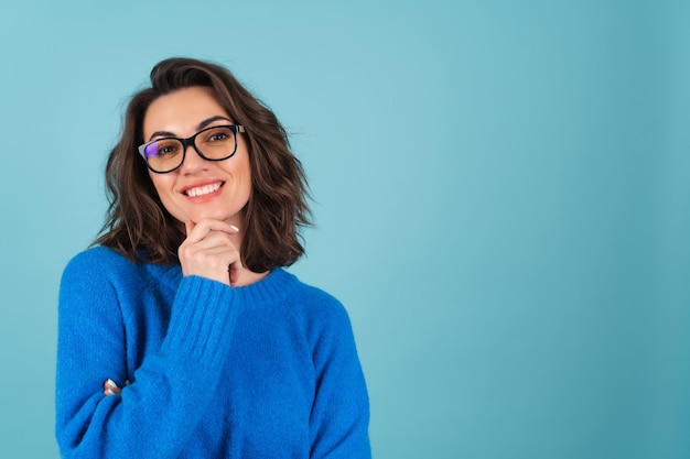 Een vrouw in een blauwe gebreide trui en natuurlijke make-up, krullend kort haar, een bril op haar ogen, kijkt bedachtzaam naar de camera, mediterend