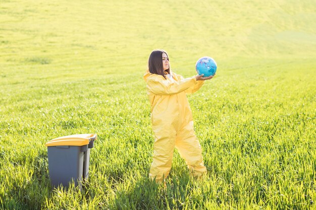 Een vrouw in een geel beschermend pak staat in het midden van een groen veld en houdt een wereldbol in haar handen naast een vuilnisbak