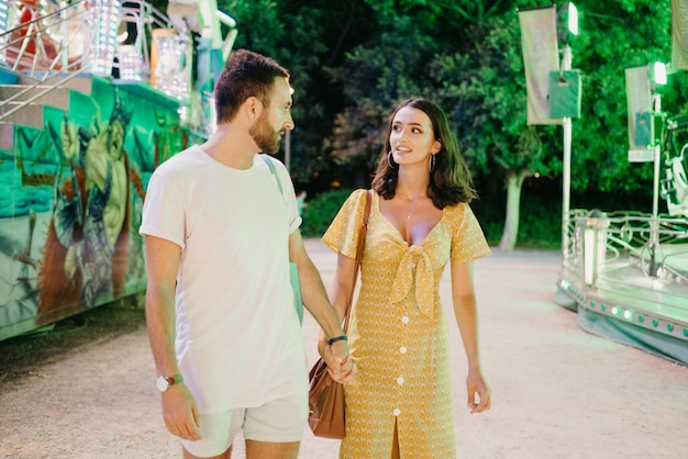 Een vrouw in een gele jurk met een decolleté en een man met een baard lopen elkaars hand vast tussen kermisattracties. Een paar geliefden op een date op de kermis in Valencia.