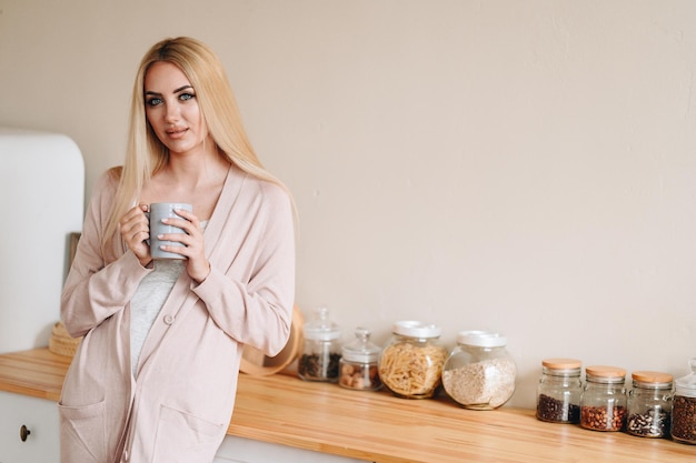 Een vrouw in een gezellige keuken drinkt koffie Beige keuken met houten tafelblad Blond in huiskleding