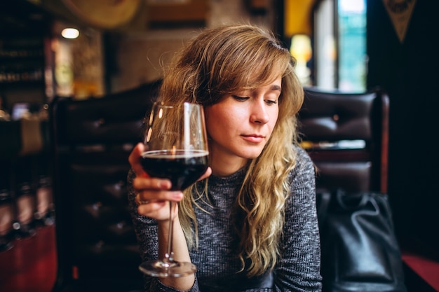 Een vrouw in een grijze top zit alleen in een restaurant met een glas wijn. romantisch meisje met blonde krullen, ontspannen in een cafe en verlegen glimlach, naar beneden te kijken.