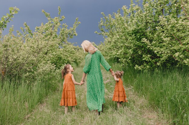 Een vrouw in een groene jurk loopt met twee kleine meisjes in een lenteappelboomgaard.