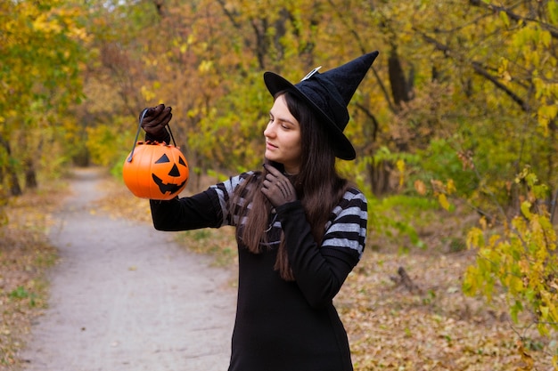 Een vrouw in een heksenkostuum houdt een pompoen met snoep vast in het herfstbos.