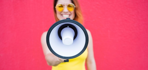 Een vrouw in een jurk met bril en oorbellen staat in profiel op een gele achtergrond en schreeuwt in een megafoon Portret van een meisje met een luidspreker Dame schreeuwt tegen een geluidsversterkingsapparaat