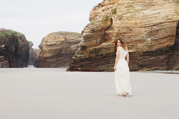 Een vrouw in een lange romantische jurk loopt blootsvoets langs een strand met kliffen Ze kijkt naar de camera