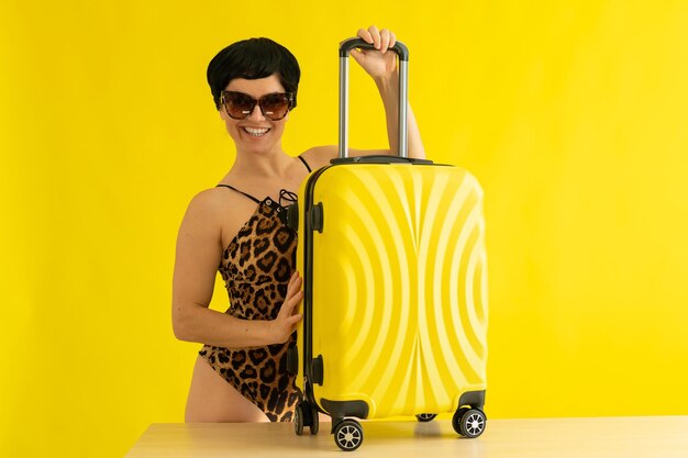 Een vrouw in een massief luipaard zwembroek en zonnebril houdt een koffer op een gele achtergrond een glimlachende brunette in een monokini heeft haar tas ingepakt en is klaar om naar de zee te reizen
