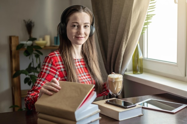 Een vrouw in een rode jurk luistert audioboek met een koptelefoon en drinkt koffie bij het raam Concept van modern technologisch onderwijs en de studie van boeken met vreugde