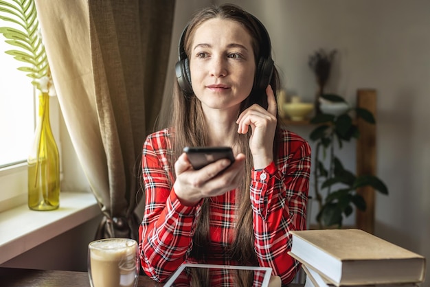 Een vrouw in een rode jurk luistert audioboek met een koptelefoon met telefoon bij het raam Het concept van modern technologisch onderwijs en de studie van boeken met vreugde