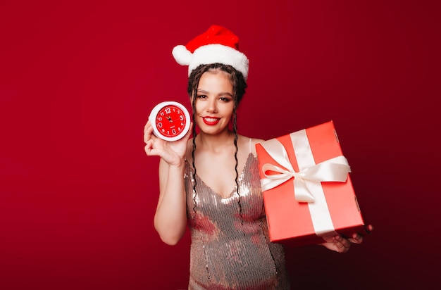 Een vrouw in een rode kerstmuts met dreadlocks houdt een geschenk op een rode achtergrond Isoleer een vrouw in een slimme jurk nieuwjaarskerst