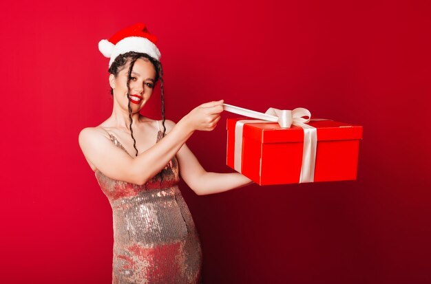 Een vrouw in een rode kerstmuts met dreadlocks houdt een geschenk op een rode achtergrond Isoleer een vrouw in een slimme jurk nieuwjaarskerst