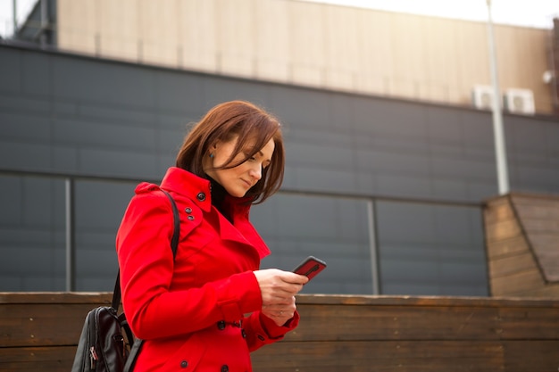 Een vrouw in een rode regenjas praat aan de telefoon in de buurt van een zakencentrum. Een brunette met een kort kapsel in de herfst of lente doet zaken, communiceert op afstand