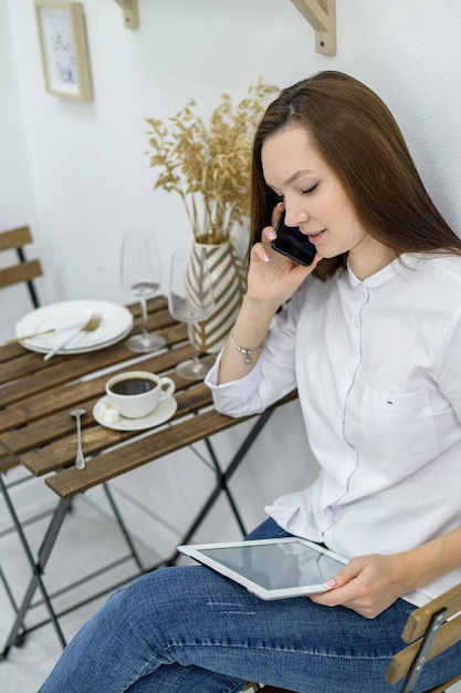 Een vrouw in een wit overhemd en spijkerbroek zit in een café met een tablet in haar handen Vrouwelijke kantoormedewerker tijdens de lunchpauze praten aan de telefoon Manager tijdens een zakelijke bijeenkomst in een restaurant drinkt koffie