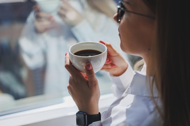 Een vrouw in een wit overhemd met geurige warme koffie in haar handen Close-up vrouw handen met een kopje americano selectieve focus