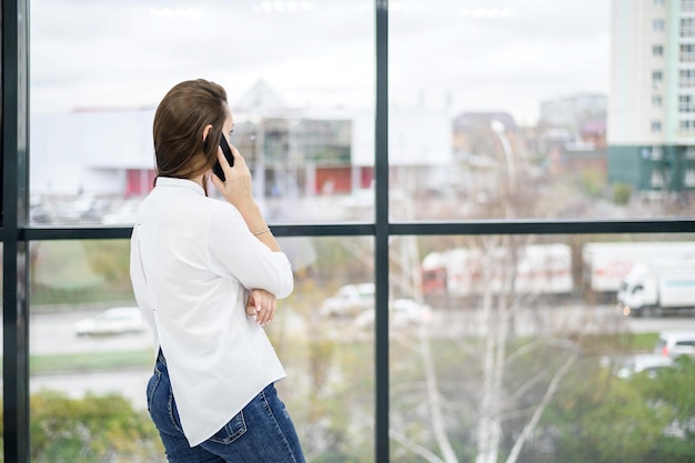 Een vrouw in een wit overhemd staat bij het raam en praat aan de telefoon Europees meisje belt op een smartphone en glimlacht