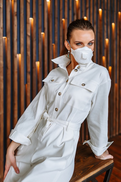 Een vrouw in een witte jumpsuit-jurk met een antiviraal masker op haar gezicht. Model zittend op een stoel