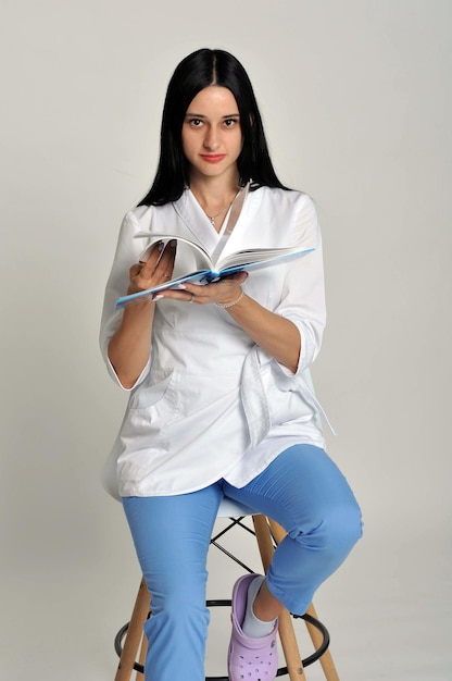 Een vrouw in een witte laboratoriumjas zit op een kruk en houdt een boek vast.