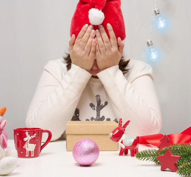 Een vrouw in een witte trui zit aan tafel en pakt cadeaus voor Kerstmis in. Houd je hoofd vast met je handen, verdriet